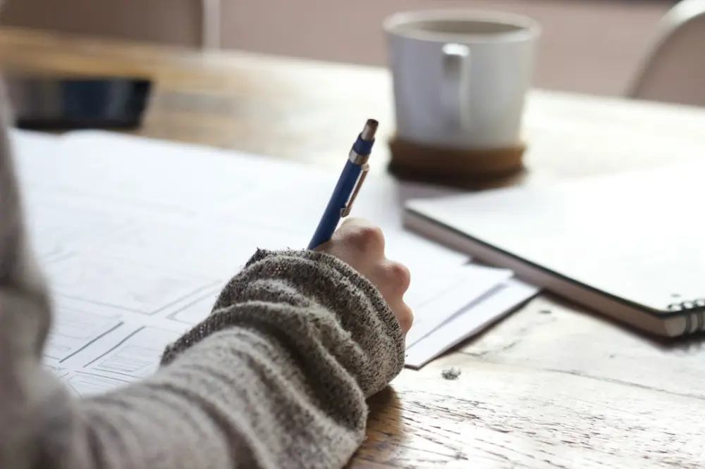 Person writing on brown wooden table near white ceramic mug, illustrating writing and learning